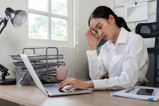 A businesswoman in a white shirt experiencing a headache while working on a laptop at her desk in a modern office setting.