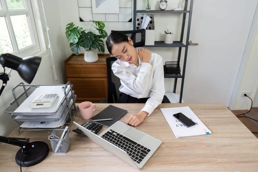 A business professional in a modern office setting experiencing neck pain while working at a desk with a laptop and office supplies.