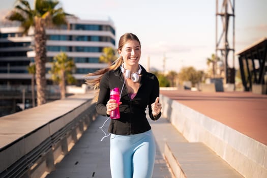 A young fitness girl runs with headphones and water bottle outdoors. Concept of workouts and healthy lifestyle.
