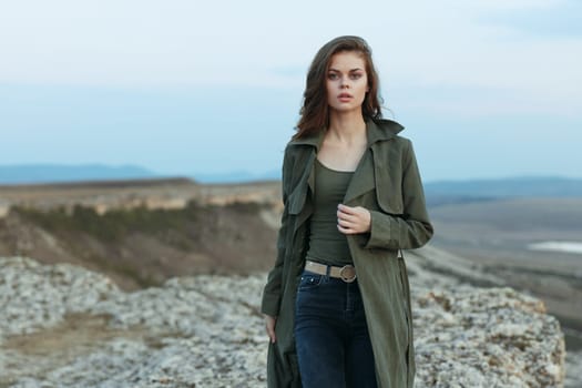 Serene woman in trench coat and jeans standing on rocky hilltop overlooking majestic mountains