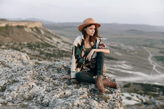 Serene woman in hat and boots sitting on rock in mountainous landscape with majestic mountains in background