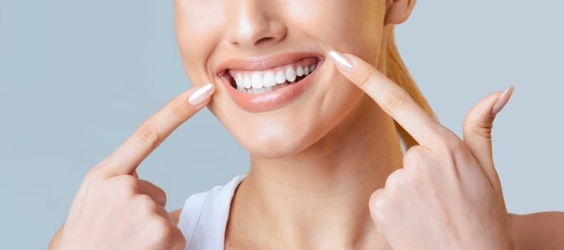 This image is a close up of a woman face. Her fingers are resting on her cheeks, and she is smiling widely, showing off her bright white teeth, cropped