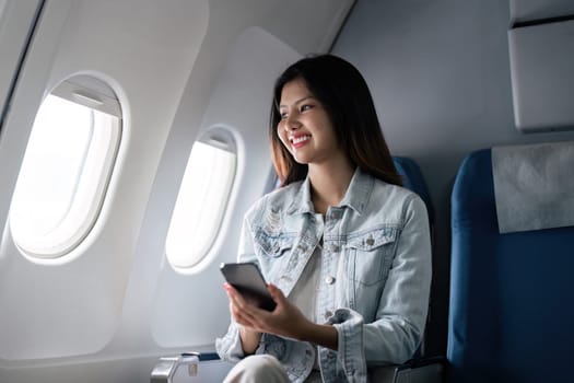 A cheerful tourist sitting in an airplane seat, holding a smartphone, and looking out the window during a flight.