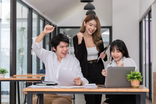 business team of three people, including a woman and two men, celebrating success at an office desk with a laptop and paperwork, bright modern office setting