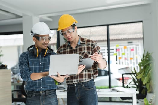 Two male engineers in a modern office, wearing safety helmets and glasses, reviewing project plans on a laptop and tablet.