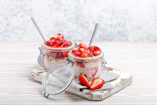 Parfait with Fresh Strawberries, Yoghurt and Crunchy Granola in Transparent Glass Mason Jars on White Rustic Wooden Background from Angle View, Healthy Breakfast or Light Summer Fruit Dessert