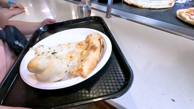 Close-up of a freshly baked pizza slice on a paper plate, placed on a black tray in a pizzeria. The background shows more pizzas being prepared, creating a warm and inviting atmosphere.