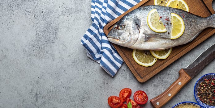 Raw fish dorado with ingredients lemon, fresh basil, cut cherry tomatoes, uncooked rice on wooden cutting board with knife on rustic stone background top view, cooking healthy fish dorado. Copy space