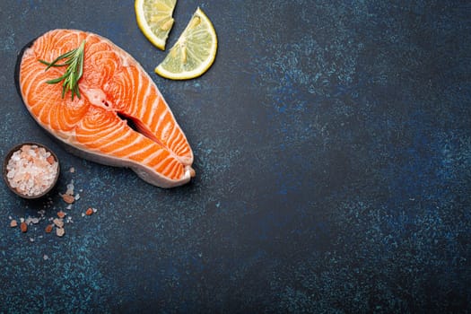 Fresh raw salmon fillet steak with rosemary on dark blue rustic stone background with rose salt and lemon top view, healthy balanced diet and nutrition with fish. Space for text