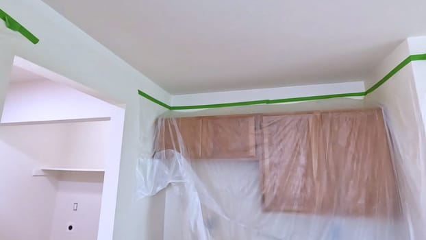 A kitchen undergoing renovation, featuring cabinets covered with plastic sheeting and masking tape along the walls and ceiling. The space is prepared for painting or further construction work.