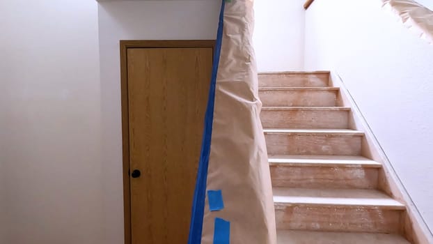 A staircase in a home undergoing renovation, featuring protective coverings and masking tape to safeguard surfaces. The area is prepared for painting or further construction work, highlighting a common home improvement project.