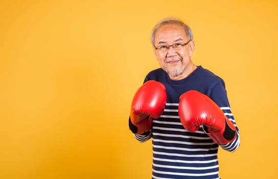 Portrait Asian old man wearing glasses wear two red boxing gloves studio shot isolated yellow background, smiling happy elderly man gray haired healthy fighter lifestyle concept