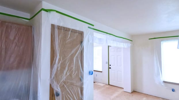 A kitchen undergoing renovation, featuring cabinets covered with plastic sheeting and masking tape along the walls and ceiling. The space is prepared for painting or further construction work.