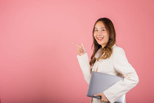 Businesswoman smiling confident smiling holding closed laptop and pointing finger to empty space, Portrait happy Asian young female person hugging close cover computer isolated on pink background