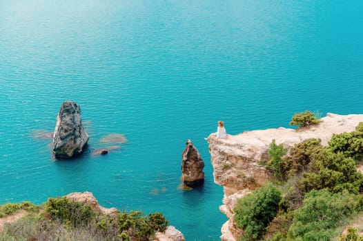 A woman sits on a rock overlooking the ocean. The water is blue and calm. The scene is peaceful and serene