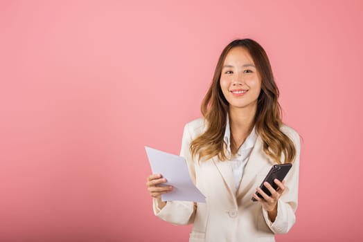 Portrait of Asian happy young woman smile holding paper calculating bills on hand and mobile phone for payment QR code, female person holding bill studio shot isolated on pink background
