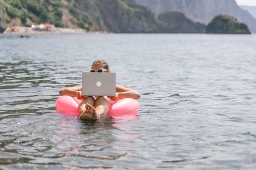 A woman is floating in a pink inflatable raft with a laptop in her hand. She is smiling and she is enjoying her time in the water