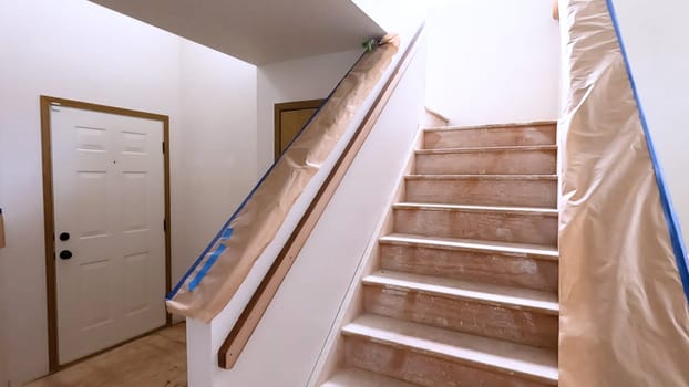 A staircase in a home undergoing renovation, featuring protective coverings and masking tape to safeguard surfaces. The area is prepared for painting or further construction work, highlighting a common home improvement project.