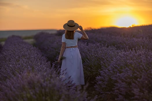 A woman is walking through a field of lavender flowers. The sun is setting in the background, casting a warm glow over the scene. The woman is wearing a straw hat and a white shirt