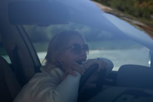 A blonde woman in a white sweater and jeans is driving. Happy woman sitting in a car with a white interior