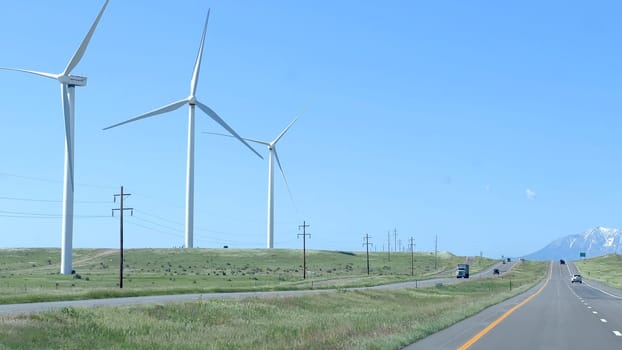 Wind turbines stand tall along a highway in Colorado, with green fields and a distant view of snow-capped mountains under a clear blue sky. Cars and trucks can be seen traveling on the road, highlighting the blend of natural and renewable energy elements.