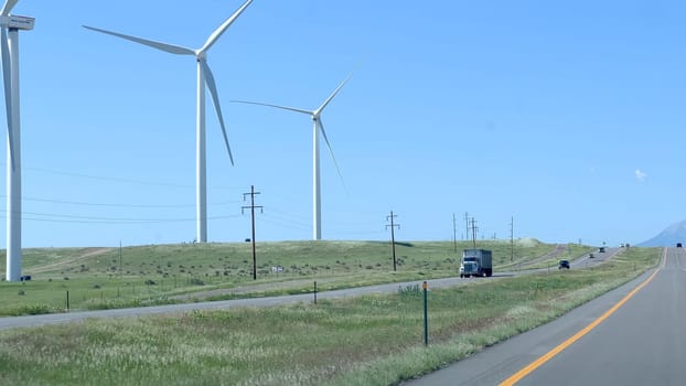 Wind turbines stand tall along a highway in Colorado, with green fields and a distant view of snow-capped mountains under a clear blue sky. Cars and trucks can be seen traveling on the road, highlighting the blend of natural and renewable energy elements.