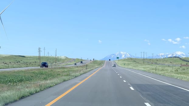 Wind turbines stand tall along a highway in Colorado, with green fields and a distant view of snow-capped mountains under a clear blue sky. Cars and trucks can be seen traveling on the road, highlighting the blend of natural and renewable energy elements.