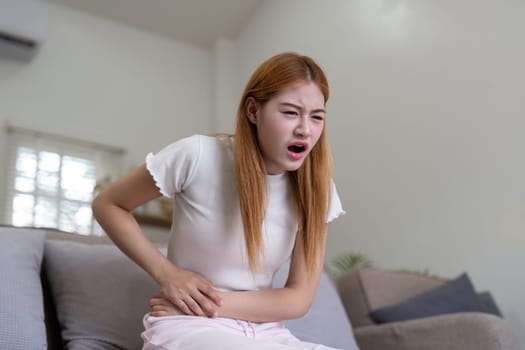 Young woman with stomach pain, sitting on a couch, holding her abdomen, expressing discomfort and pain.