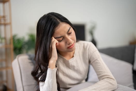 A young woman sitting on a couch, holding her head in pain, experiencing a headache and migraine, feeling sick and unwell.