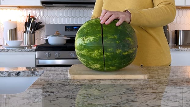 A woman in a yellow sweater slices a fresh watermelon on a cutting board in a modern kitchen. The kitchen features white cabinetry, a hexagonal tile backsplash, and various cooking utensils on the countertop.
