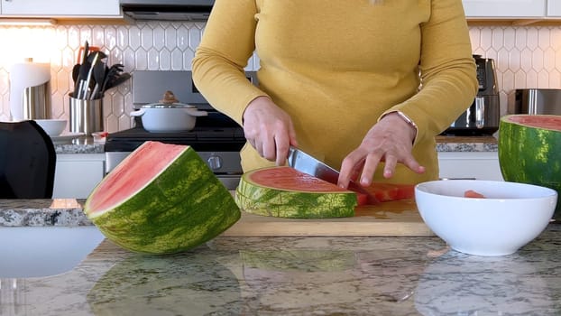 A woman in a yellow sweater slices a fresh watermelon on a cutting board in a modern kitchen. The kitchen features white cabinetry, a hexagonal tile backsplash, and various cooking utensils on the countertop.