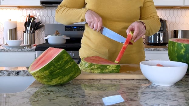 A woman in a yellow sweater slices a fresh watermelon on a cutting board in a modern kitchen. The kitchen features white cabinetry, a hexagonal tile backsplash, and various cooking utensils on the countertop.