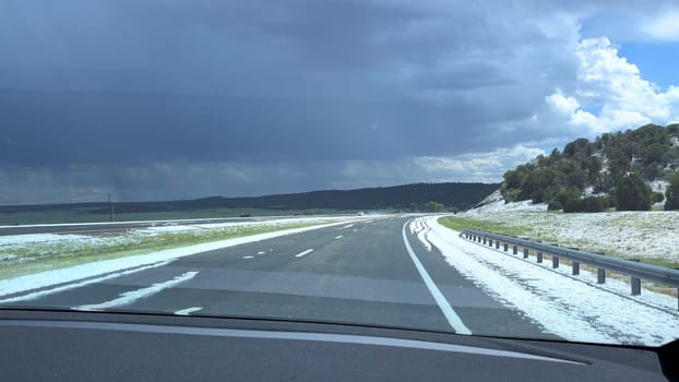 A striking image showcasing a hail-covered road after a storm in Colorado. The aftermath of the storm leaves the road and surrounding landscape blanketed in white hail, with dark storm clouds lingering in the sky.