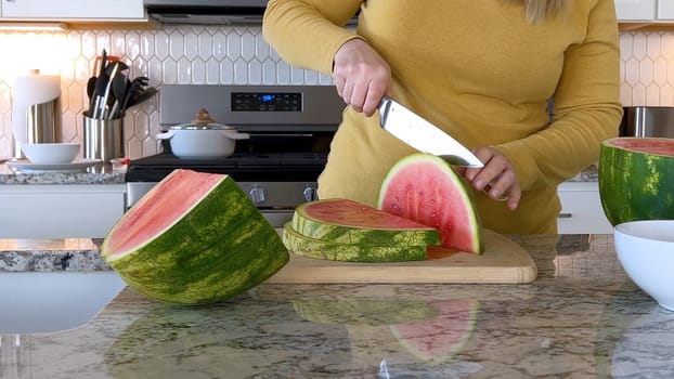 A woman in a yellow sweater slices a fresh watermelon on a cutting board in a modern kitchen. The kitchen features white cabinetry, a hexagonal tile backsplash, and various cooking utensils on the countertop.