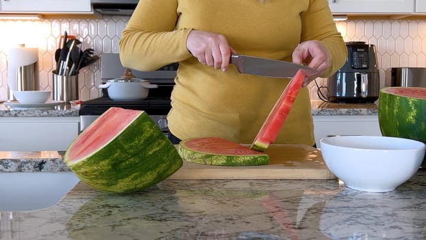 A woman in a yellow sweater slices a fresh watermelon on a cutting board in a modern kitchen. The kitchen features white cabinetry, a hexagonal tile backsplash, and various cooking utensils on the countertop.