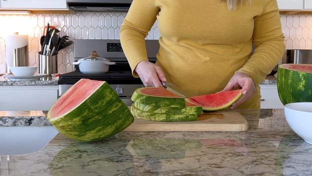 A woman in a yellow sweater slices a fresh watermelon on a cutting board in a modern kitchen. The kitchen features white cabinetry, a hexagonal tile backsplash, and various cooking utensils on the countertop.