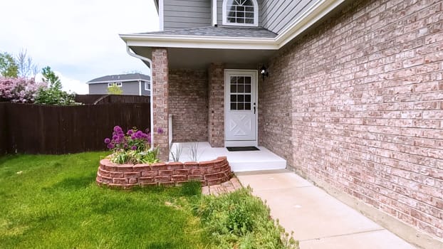 The front porch of a charming brick house featuring a white door and a well-maintained garden bed with blooming purple flowers. The neatly trimmed lawn and inviting entryway enhance the home curb appeal.