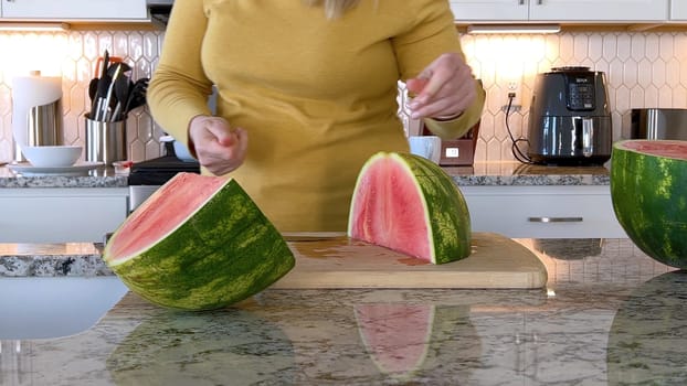 A woman in a yellow sweater slices a fresh watermelon on a cutting board in a modern kitchen. The kitchen features white cabinetry, a hexagonal tile backsplash, and various cooking utensils on the countertop.