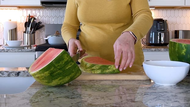 A woman in a yellow sweater slices a fresh watermelon on a cutting board in a modern kitchen. The kitchen features white cabinetry, a hexagonal tile backsplash, and various cooking utensils on the countertop.