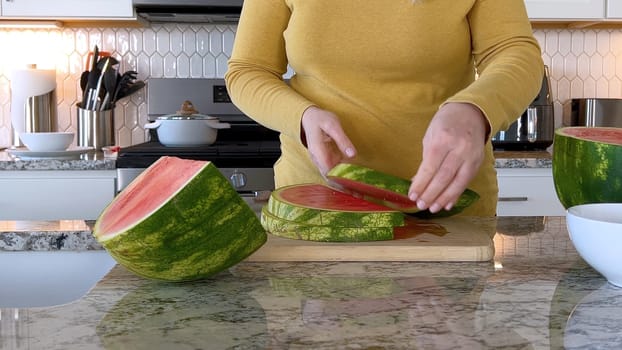 A woman in a yellow sweater slices a fresh watermelon on a cutting board in a modern kitchen. The kitchen features white cabinetry, a hexagonal tile backsplash, and various cooking utensils on the countertop.