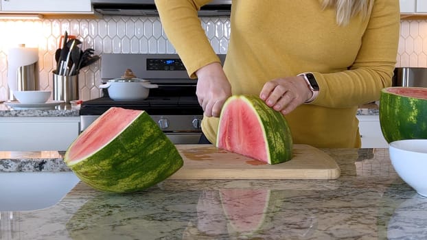 A woman in a yellow sweater slices a fresh watermelon on a cutting board in a modern kitchen. The kitchen features white cabinetry, a hexagonal tile backsplash, and various cooking utensils on the countertop.