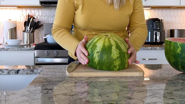 A woman in a yellow sweater slices a fresh watermelon on a cutting board in a modern kitchen. The kitchen features white cabinetry, a hexagonal tile backsplash, and various cooking utensils on the countertop.