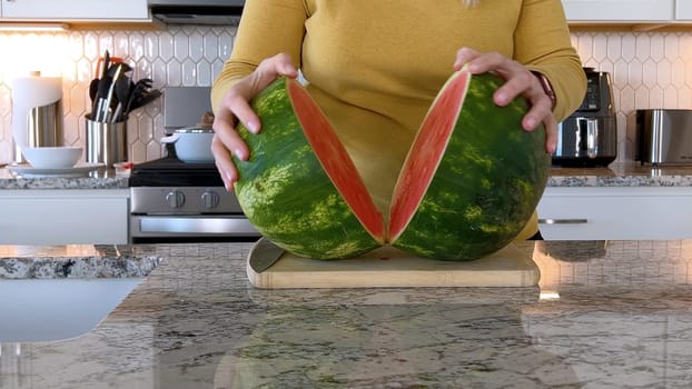 A woman in a yellow sweater slices a fresh watermelon on a cutting board in a modern kitchen. The kitchen features white cabinetry, a hexagonal tile backsplash, and various cooking utensils on the countertop.