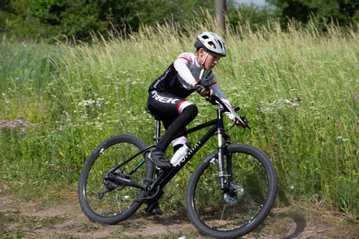 Kursk, Russia, June 15, 2024: Child cyclist on cross bike enters turn on rough terrain with high grass, mountain bike competition