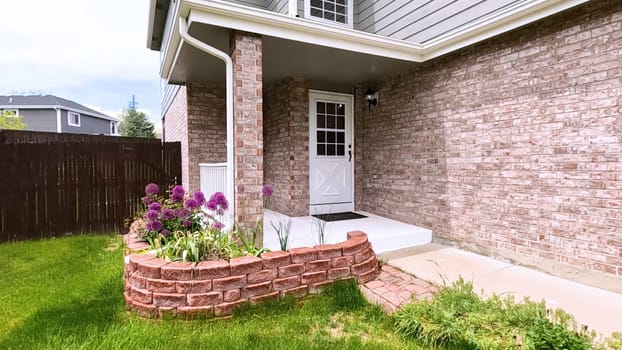 The front porch of a charming brick house featuring a white door and a well-maintained garden bed with blooming purple flowers. The neatly trimmed lawn and inviting entryway enhance the home curb appeal.