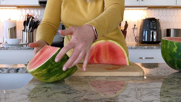 A woman in a yellow sweater slices a fresh watermelon on a cutting board in a modern kitchen. The kitchen features white cabinetry, a hexagonal tile backsplash, and various cooking utensils on the countertop.