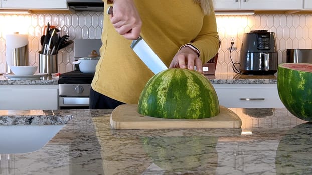 A woman in a yellow sweater slices a fresh watermelon on a cutting board in a modern kitchen. The kitchen features white cabinetry, a hexagonal tile backsplash, and various cooking utensils on the countertop.