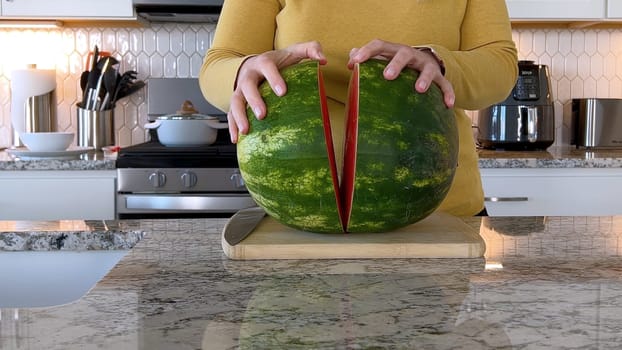 A woman in a yellow sweater slices a fresh watermelon on a cutting board in a modern kitchen. The kitchen features white cabinetry, a hexagonal tile backsplash, and various cooking utensils on the countertop.