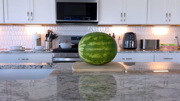 Fresh watermelon halves are placed on a cutting board in a modern kitchen. The kitchen features white cabinetry, a hexagonal tile backsplash, and various cooking utensils and appliances on the countertop.