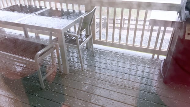 Image capturing a hail storm on a wooden deck, highlighting the impact of hail on patio furniture and the deck surface. Hailstones are visible scattered across the wet deck, with patio furniture in the background.